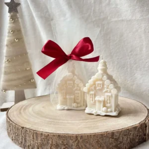 Dwie świece sojowe w kształcie domków świątecznych ustawione obok siebie, jedna zapakowana w tiul.