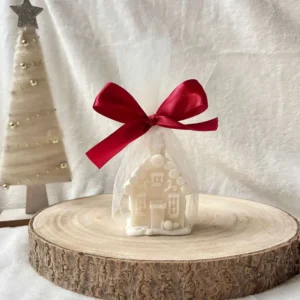 Świeca sojowa w kształcie domku świątecznego, zapakowana w biały tiul z czerwoną wstążką.