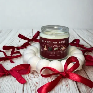 Świeca sojowa Plany na dziś ustawiona na drewnianym tle wśród czerwonych wstążek, tworząca ciepły i elegancki klimat.